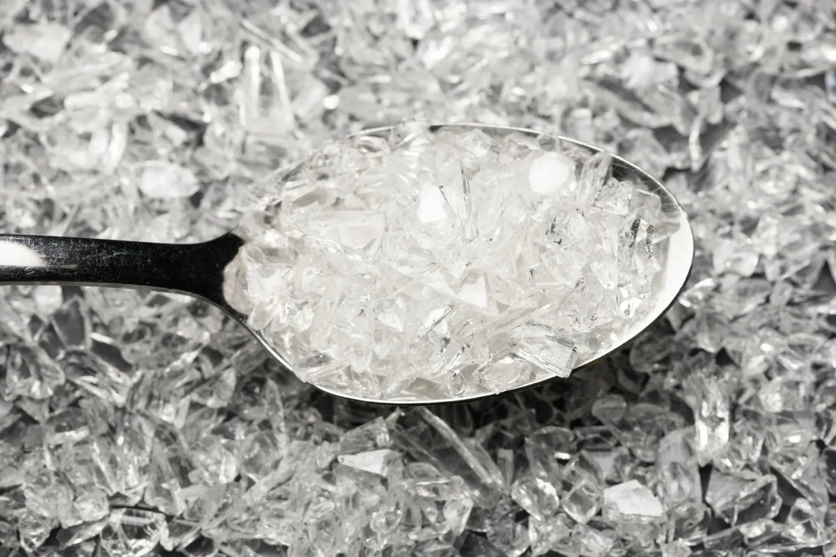 A spoonful of glass frit held over a pile of the same glass frit in an off white colour.
