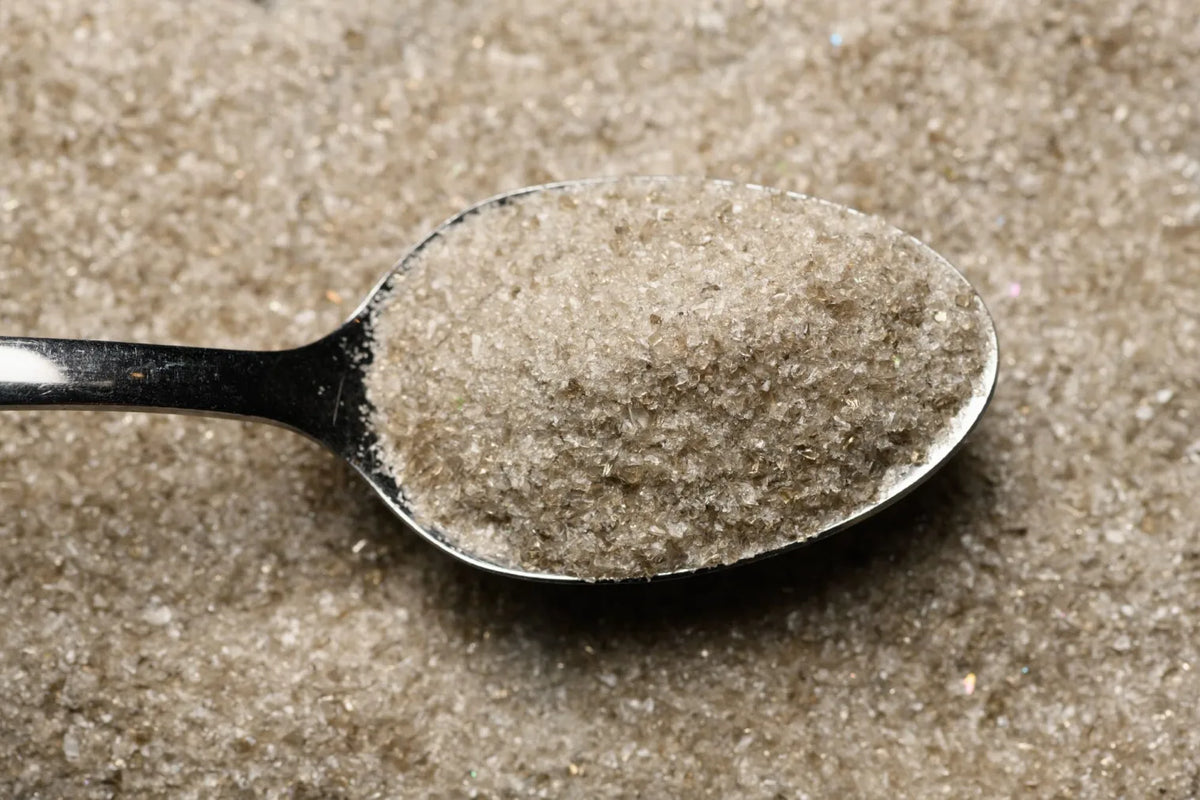 A close-up of a metal spoon filled with glass frit, set against a background of matching granules.