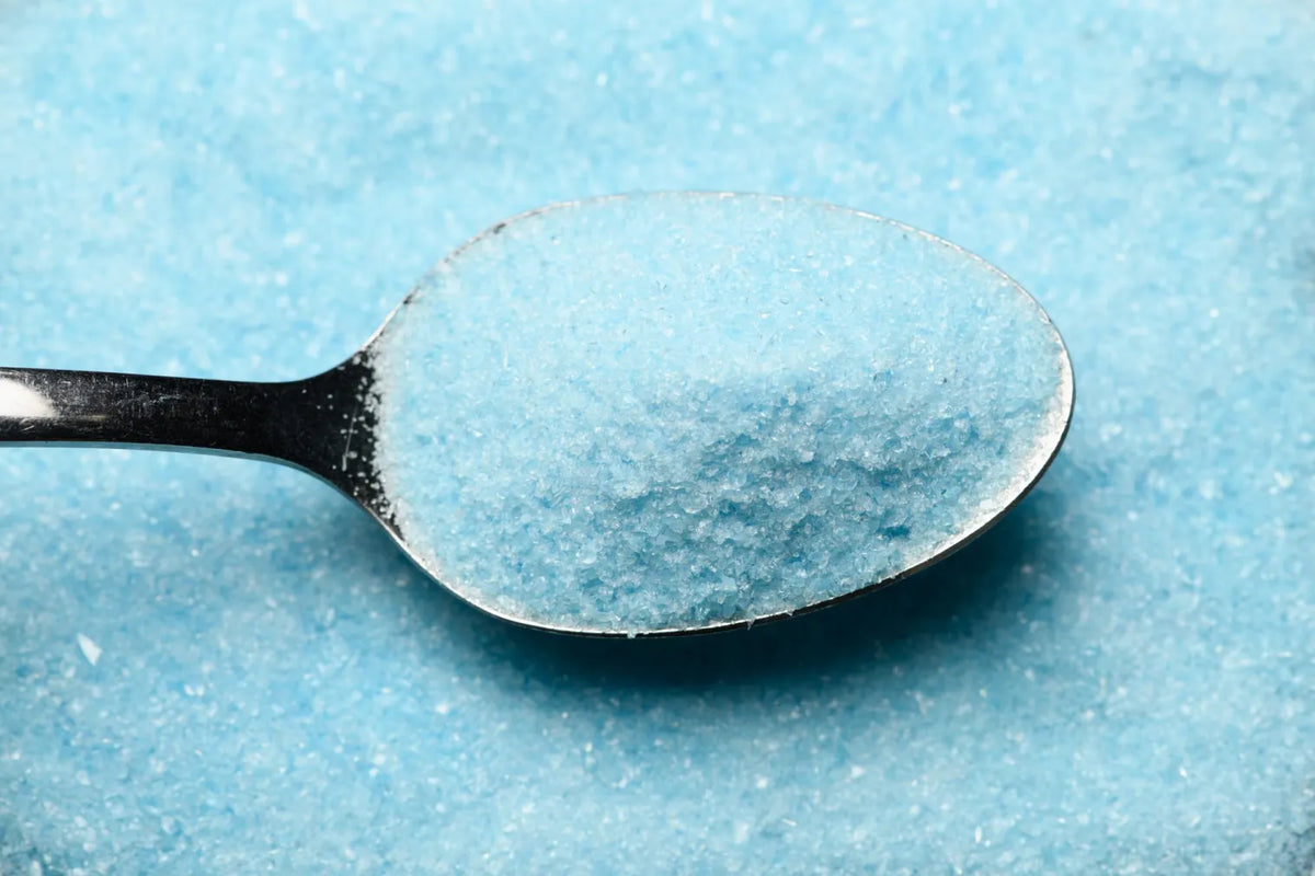 A close-up of a metal spoon filled with light blue granulated powder, set against a textured light blue background.