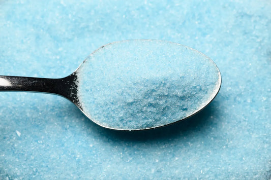 A close-up of a metal spoon filled with light blue granulated powder, set against a textured light blue background.