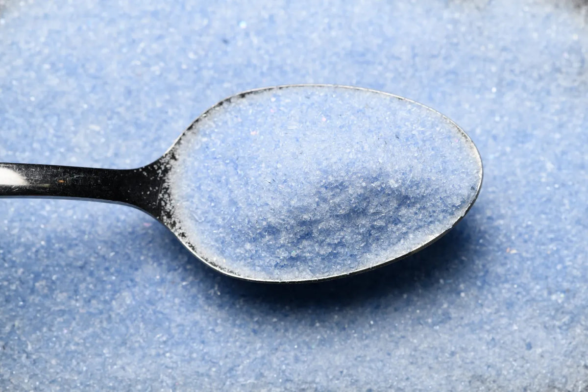 A close-up of a metal spoon filled with fine light blue powder or granules, set against a matching textured blue background.